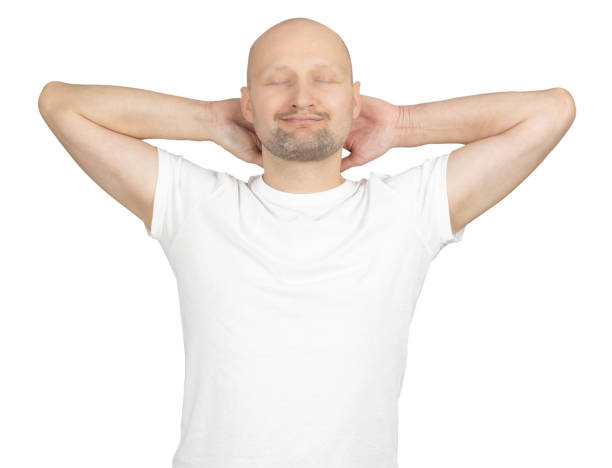 A Caucasian bald man wearing a white t-shirt is standing against a plain background. He has his hands placed behind his head and eyes closed, displaying a relaxed and content expression. The image conveys a sense of calm, ease, and comfort.