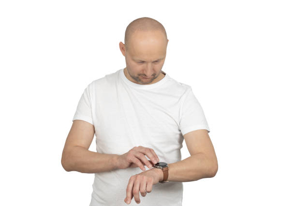 A bald Caucasian man with light stubble is wearing a white t-shirt and looking down at his smartwatch. His expression is focused, suggesting he is checking the time or notifications. The image conveys a concept of time management, technology, and daily routine.