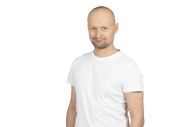 A bald Caucasian man with a short beard is wearing a white t-shirt and giving a subtle, charming smile while looking directly at the camera. His confident and relaxed posture conveys warmth and friendliness. The neutral background keeps the focus on his expression.