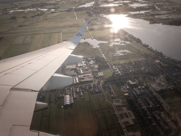 SCHIPOL, NETHERLANDS - AUGUST 12, 2024: image of a KLM plane wing gliding over the Dutch countryside, offering a sweeping panorama of quintessential Netherlands polder land. Taken from a window seat, the photo highlights neat farmland parcels, intricate canals, and shimmering waterways that define this historic region near Amsterdam. The soft light casts a warm glow on the terrain, emphasizing the harmonious balance between agriculture and water management in the Netherlands. Ideal for travel and features.