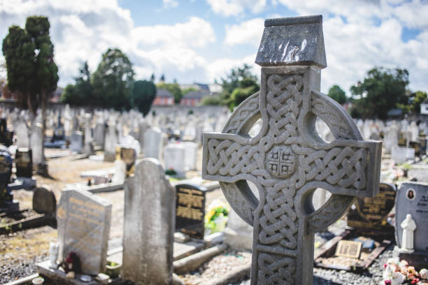 A detailed Celtic cross gravestone with intricate carvings stands prominently at Glasnevin Cemetery in Dublin, Ireland, surrounded by tombstones under a bright sky.