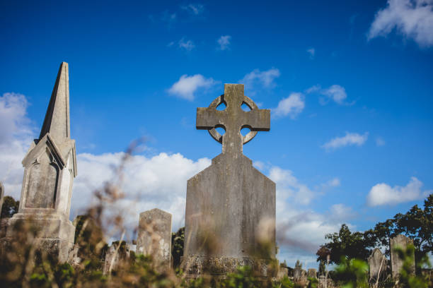 A weathered Celtic cross tombstone stands against a vibrant blue sky in Glasnevin Cemetery, Dublin, Ireland.
