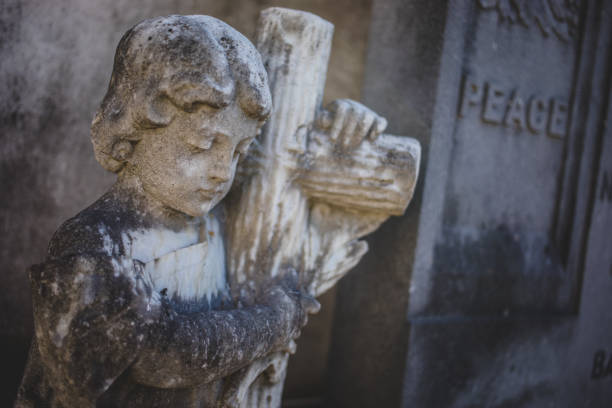 A mature woman, dressed in black mourning attire with a headscarf and black veil, standing solemnly in front of a tombstone at a cemetery. The scene conveys a sense of grief and respect, capturing the tradition of mourning and reflection at a final resting place. The soft lighting and somber expression evoke emotions of loss, remembrance, and reverence.