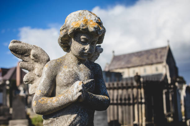 Dublin, Ireland - September 21, 2018: A weathered angel statue with folded hands stands in Glasnevin Cemetery, with a historic church in the background.