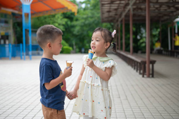 Young boy and girl holding hands and eating ice creams on a park pathway. The boy and girl are walking hand in hand on a park's paved path, each enjoying an ice cream cone, displaying a moment of friendship and joy in an outdoor setting with visible structures in the background. 3 year old boy and 4 year old girl ice cream time asian little chinese girls eating ice cream stock pictures, royalty-free photos & images