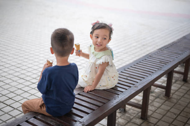 This captivating image features a young girl and a boy sitting comfortably on a bench while playfully enjoying her ice cream, radiating joy and innocence, set against a lush and vibrant park background.