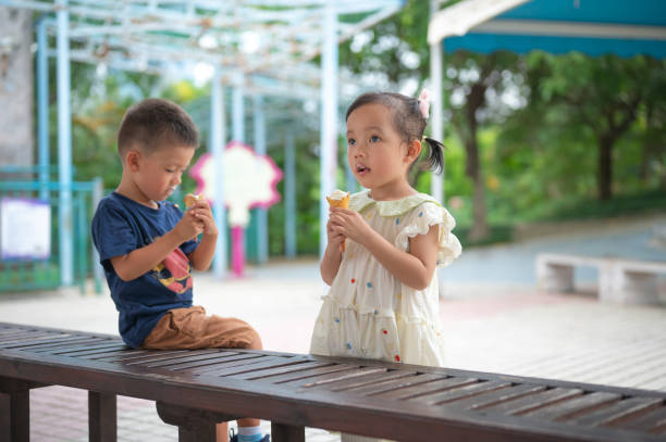 Playful Children Savoring Ice Cream in Park Two playful children enjoying their ice cream while sitting on a bench, showcasing the joyful essence of childhood, friendship, and outdoor fun in a park environment. asian little chinese girls eating ice cream stock pictures, royalty-free photos & images