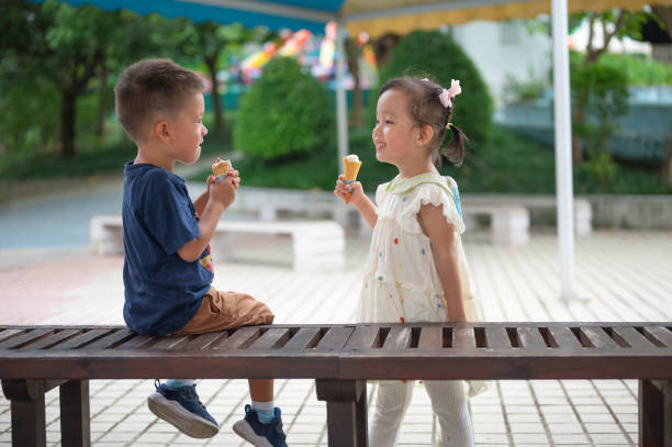 Children Enjoying Ice Cream in a Park Setting A cheerful scene featuring two young children sharing a delightful moment while savoring ice cream in a vibrant park, surrounded by greenery and a friendly atmosphere that promotes joy and friendship. asian little chinese girls eating ice cream stock pictures, royalty-free photos & images