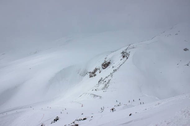 Snow is lifted by strong winds across an open mountain slope, revealing a vast panorama of distant peaks and clouds in a cold, silent high-altitude environment.