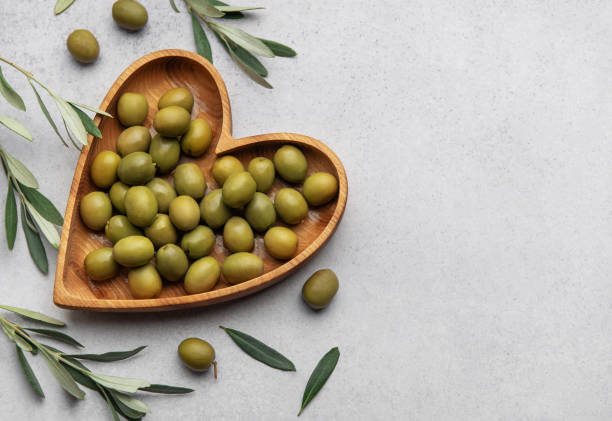 istock Fresh green olives displayed on wooden heart shaped bowl with olive branches 2199294369