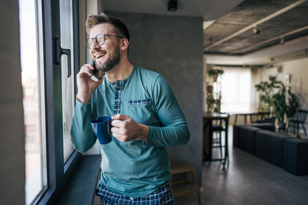 istock Smiling man in pajamas drinking coffee and talking on phone by window 2199323385