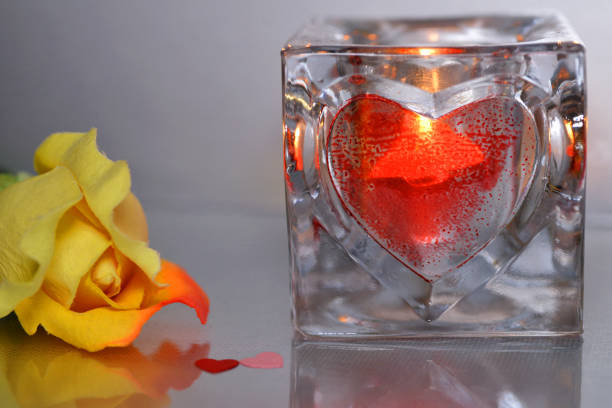 A close-up shot of a red heart-shaped candle inside a glass holder, glowing warmly against a dimly lit background. The flickering light enhances the sense of romance and passion, making this a perfect image for intimate and emotional themes.