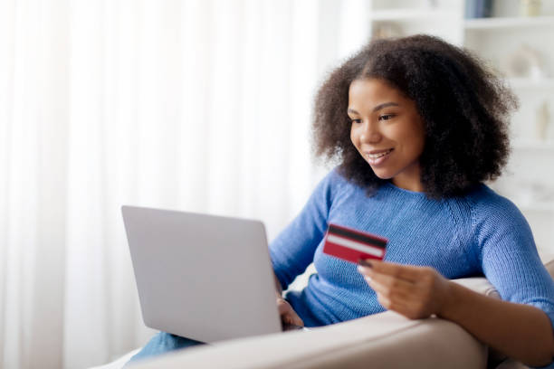istock Online Shopping. Happy Black Woman Using Pc Laptop And Credit Card Making Payment Using Internet Banking Sitting On Sofa At Home. Female Customer Paying With Bankcard Ordering Delivery On Website 2199335033