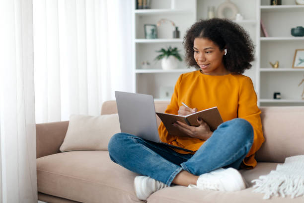 istock Portrait of smiling woman sitting on sofa, using laptop and writing in notebook, taking notes, watching tutorial, lecture or webinar, studying online at home looking at screen, free copy space 2199337139
