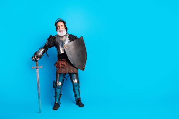istock Elderly man in medieval knight costume posing against blue background, holding shield and sword with expression of bravery and humor 2199337315