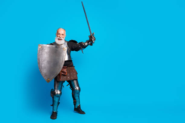 An older gentleman in full metal medieval armor, holding a sword and shield, set against a vibrant blue background, evokes themes of chivalry and playful costume.