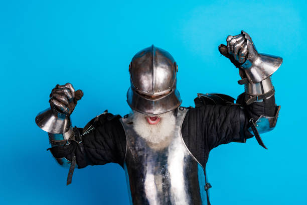 A bearded older man in full medieval knight armor joyfully celebrating against a blue studio background, showcasing historical charm with vibrant energy.