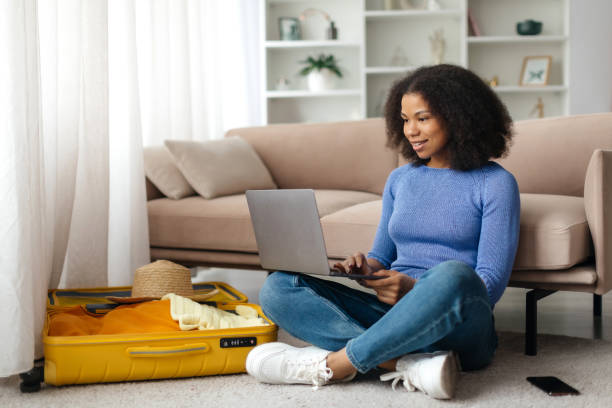 istock Excited african lady using laptop and talking on phone while searching summer trip, preparing for holidays. Happy woman surfing internet for weekend travel at home 2199339907