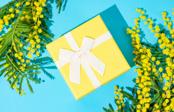 istock Yellow gift box on blue background with mimosa flowers and sunlight. 2199340253