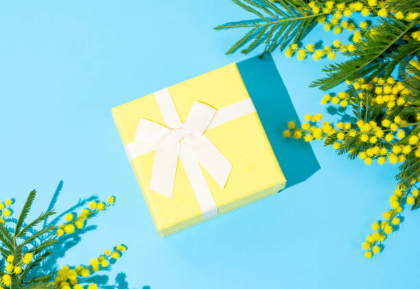 istock Yellow gift box on blue background with mimosa flowers and sunlight. 2199340259