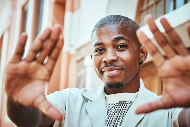 Smiling young man standing outdoors and creatively framing with his hands. Captures a cheerful moment on a sunny day in an urban setting, showcasing self-expression and connection to the environment.