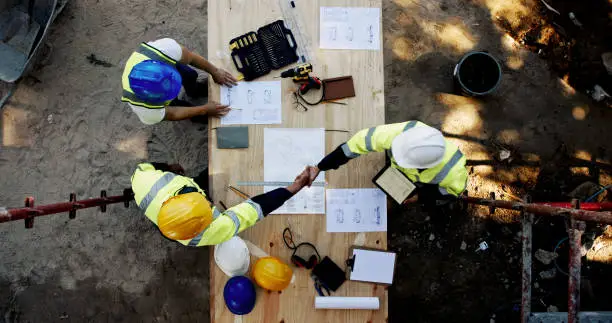 Blueprint, architecture and men at table shaking hands for collaboration, agreement and inspection from above. Construction, building team and contractor on site with floor plan, handshake and deal Blueprint, architecture and men at table shaking hands for collaboration, agreement and inspection from above. Construction, building team and contractor on site with floor plan, handshake and deal