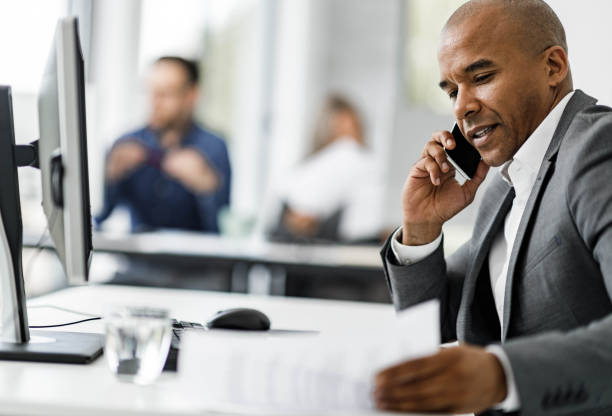 istock Smiling black businessman reading notes while talking on cell phone in the office. 2199355367
