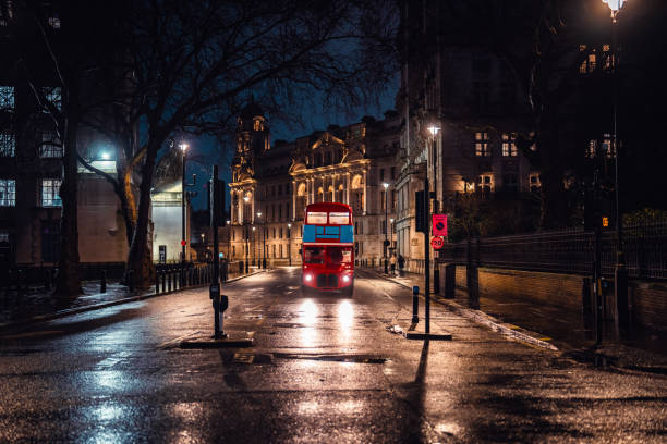 Classic red double-decker bus drives through a dimly lit london street, showcasing historic architecture