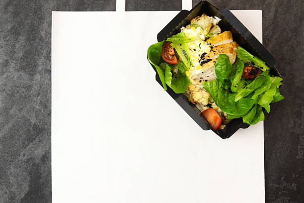 Salad take away. Healthy salad with different lettuce, corn, cauliflowers and chicken in black paper box on paper eco bag on gray background. Copy space. Healthy restaurant food delivery concept.