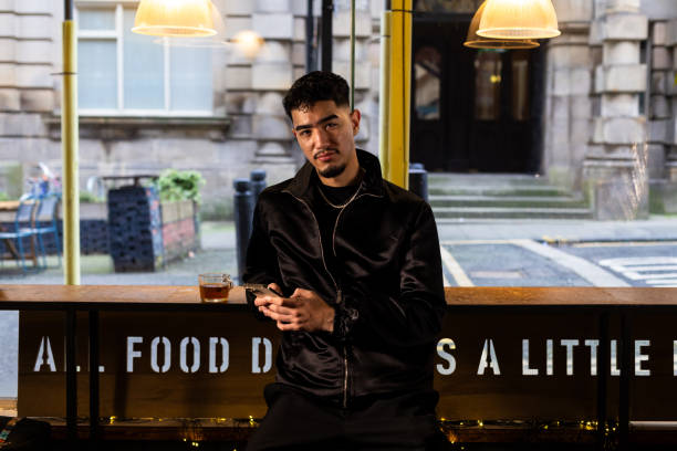Medium shot of a young man wearing a black jacket while sitting down by himself in a coffee shop in Newcastle, Northeast of England. The man is looking directly at the camera while holding a mobile phone. He has a cup of tea next to him.Videos are available similar to this scenario.
