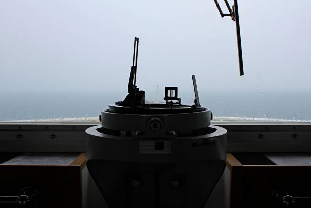Close-up of a gyrocompass repeater on the ship's bridge, a crucial navigational instrument. This image highlights the precision and detail of maritime equipment, essential for seafaring and nautical navigation.  The compass points towards the viewer, with a blurred, rainy window and ocean in the background, suggesting a journey or voyage.