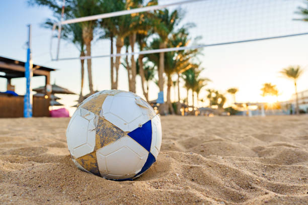 A weathered soccer ball sits on the soft sand of a beach. In the background, palm trees line the area as the sun sets, creating a warm, relaxing atmosphere perfect for sports.