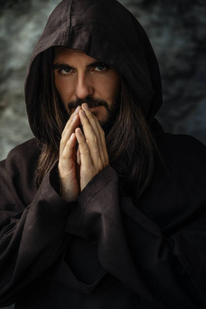 Bearded man with long hair wearing a hooded robe, hands clasped in prayer, standing against a dark, rocky background, evoking spiritual reflection.