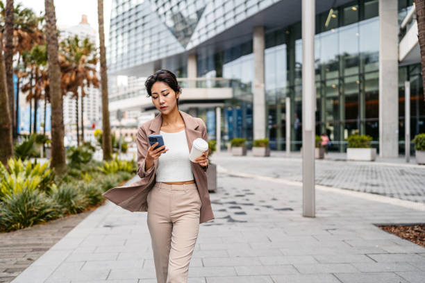 istock Young Asian Businesswoman Using Phone And Drinking Coffee To Go While Walking In Darling Harbor In Sydney In Australia 2199391047