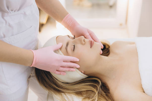 Cosmetic professional performing facial massage on relaxed blonde client following aesthetic procedure in medical spa setting Beautician wearing pink gloves massaging face of young blonde woman lying on couch with closed eyes after injection treatment in beauty clinic blonde massage stock pictures, royalty-free photos & images