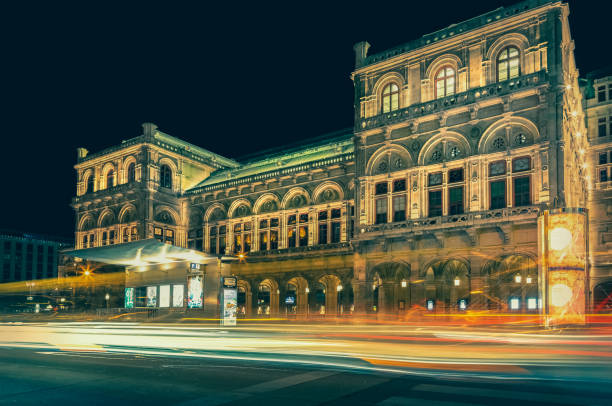 Illuminated Vienna State Opera at night, featuring vibrant light trails from passing cars and a ticket booth in the foreground, capturing the essence of Vienna's cultural heritage