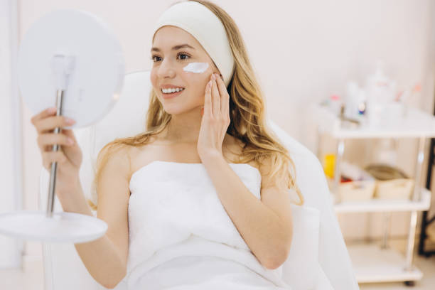 istock Smiling beautician applying moisturizing cream on her face, looking at a mirror in a beauty salon 2199396840