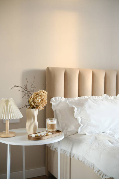 istock A calm and serene morning bedroom scene displays a chic bedside white round table complete with an elegant dry branches and flowers, a refreshing coffee drink, wooden tray with cosmetic accessories. 2199403373