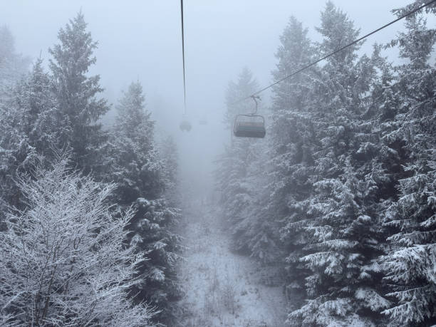 A ski lift glides through a breathtaking winter landscape, suspended above a snow-covered alpine forest shrouded in thick fog. The frost-covered trees and misty atmosphere create a peaceful yet adventurous scene, emphasizing the isolation and beauty of mountain travel in winter conditions. The empty chairlifts disappearing into the mist add a sense of mystery and remoteness.