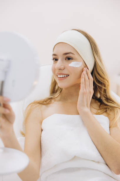 istock Skincare routine showing woman wearing headband, applying facial moisturizer while checking mirror during spa treatment session 2199403735