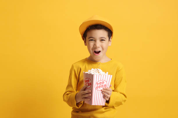 Excited kid with cap and holding a box of popcorn and posing against a yellow background