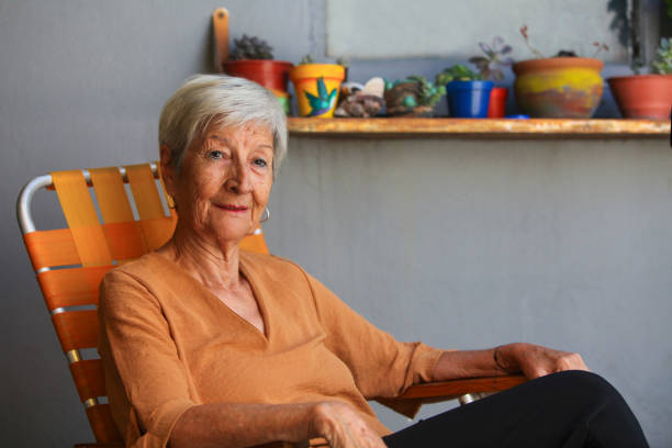 portrait of latino senior woman sitting in her patio looking at camera with pots of flowers in background