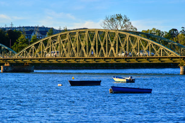 Traffic crossing metal bridge over blue river with moored boats on sunny day. Colindres, cantabria, spain