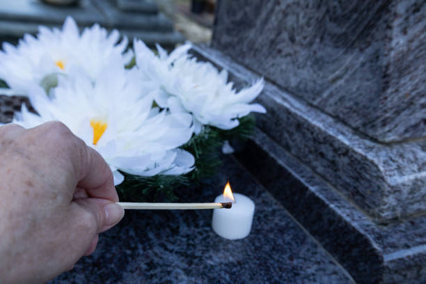 A close-up of a hand using a matchstick to light a small white candle on a gravestone. White artificial flowers are placed nearby, symbolizing remembrance and tribute at a cemetery.