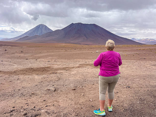 Woman in pink jacket standing in arid desert. Vast open terrain with hills, blue sky, and clouds. Solo travel and exploration in remote natural scenery.