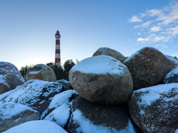 Osinovets Lighthouse rises in the background behind large snow-covered boulders, creating a dramatic contrast against the clear evening sky. The golden glow of the setting sun highlights the winter landscape, emphasizing the rugged texture of the rocks and the lighthouse as a navigational landmark on Lake Ladoga