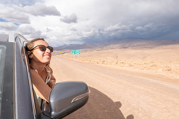 Smiling woman leans out car window enjoying desert scenery. Joyful traveler exploring open road under dramatic sky. Road trip adventure through arid landscape.