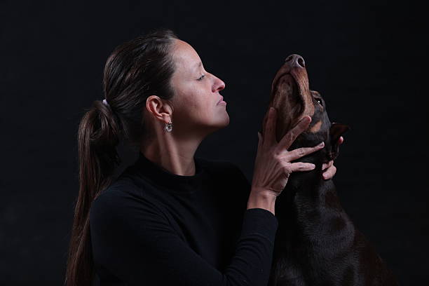 young woman in a black turtleneck with long hair looks closely at a Doberman dog with its head raised, on a black background, photographed in a studio