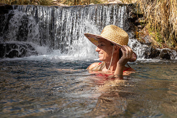 A mature woman splashes in the cool water of a lake, basking in the summer sunlight. She enjoys the refreshing feel of the water while surrounded by vibrant greenery and scenic beauty.