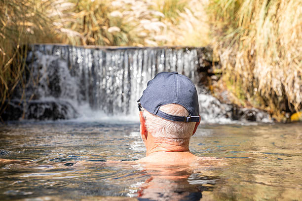 Man wearing cap, enjoying a soak in a natural hot spring. Peaceful outdoor setting with a small waterfall. Tranquil retreat surrounded by nature.